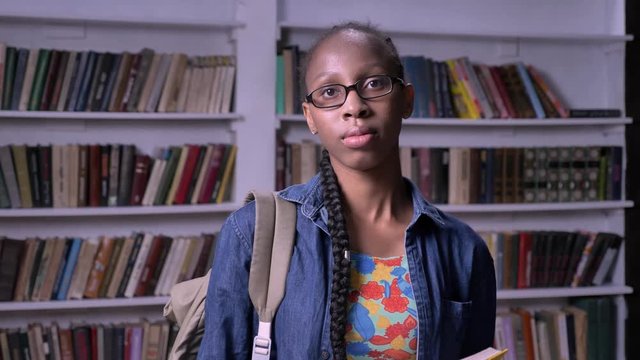 Young pretty african american woman in glasses reading book in library, looking at camera, serious and concentrated, bookshelves background