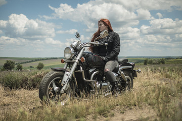 Obraz na płótnie Canvas The red-haired biker girl is sitting on a motorcycle. field of meadow and clouds
