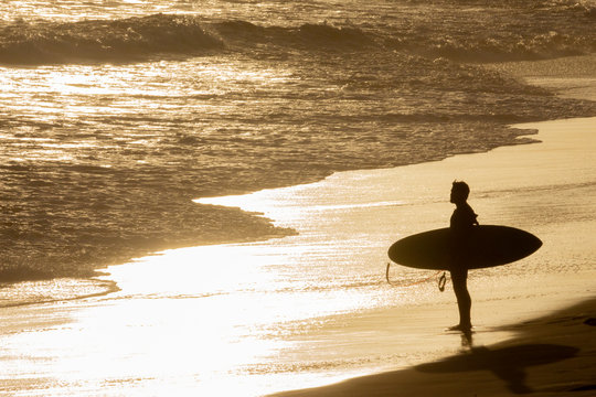 Silhouetted, isolated surfer standing on golden sand before sunwashed waves