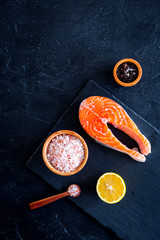 Raw red fish steak. Peiece of fresh salmon on cutting board near sea salt, pepper, lemon slices on black background top view copy space