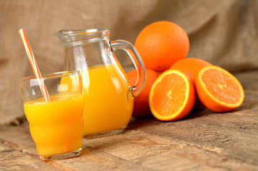 Fototapeta na wymiar Glass with orange juice and straw, jug with fresh juice and pile of oranges in the background on wooden table