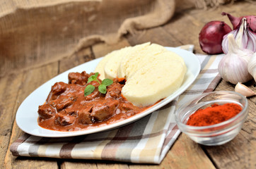 Pork goulash meat with dumplings on white plate, cutlery, garlic, onion, pepper, tablecloth in the background - typical Czech food