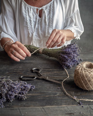 Female florist holding bunch of lavender tied by twine. Working table with lavenders, old scissors...