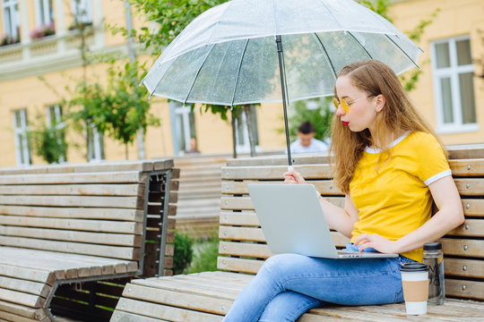 Hipster Girl With Red Lips In Yellow T-shirt Sitting On Bench Under Umbrella Outside Of Cafe With Coffee Disposable Cup And Working Laptop.