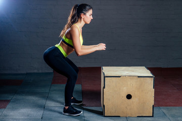 a young woman in a green sports bra and black pants jumps on a box against a backdrop of black...