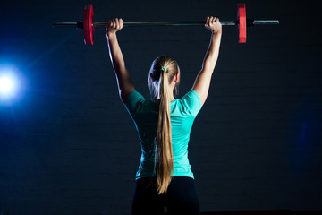 young woman in turquoise t-shirt and black pants doing exercises with a red barbell on a black...
