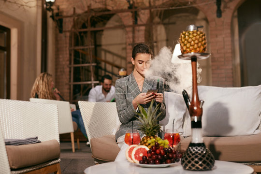 Woman With Phone Smoking Fruit Hookah In Shisha Bar