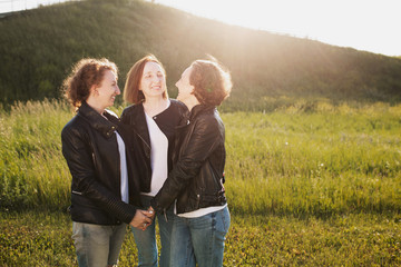 Concept of youth and maturity. Nice mother hugs her adult daughters dressed in the same clothes with a white T-shirt in jeans and leather jackets against the backdrop of a green hill on a warm sunny d