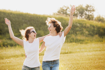 Beautiful young girls sisters in sunglasses the same clothes in white t-shirts and jeans hug and laugh on the green field outside the city