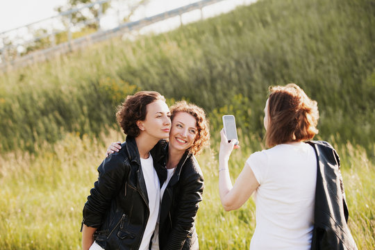 Pretty slim mom takes pictures on smartphone her adult positive beautiful daughters walking in the park in the same clothes and white T-shirts in jeans and leather jackets