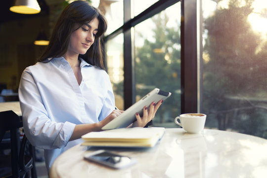 Cheerful young beautiful woman using her touchpad. Beautiful cute young businesswoman in the cafe using digital tablet and drinking coffee.