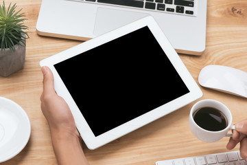 Mock up image of a hand holding black tablet pc with white blank screen and coffee cup on wooden table background