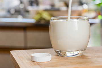 Hand pouring fresh kefir probiotik drink from white bottle into clear glass on kitchen table