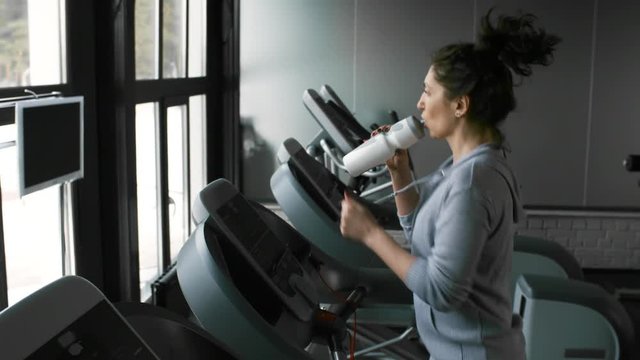 Athletic young woman taking bottle and drinking water when exercising on treadmill during workout at the gym