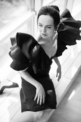 The young bewitching brunette lady in a black dress sitting on the floor indoor and looking aside