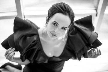 Portrait of the young beautiful brunette lady in a black dress who is sitting near the window indoors