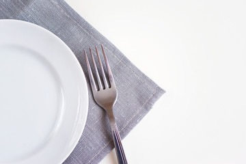 Empty white plate on a gray napkin with fork on white table. Table setting, preparation for food.