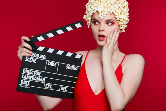 Closeup of a lady in a popcorn wig, wearing red eye shadow and lip gloss. The girl posing on the burgundy background, holding the clapperboard, looking to the side with anxious expression on her face.