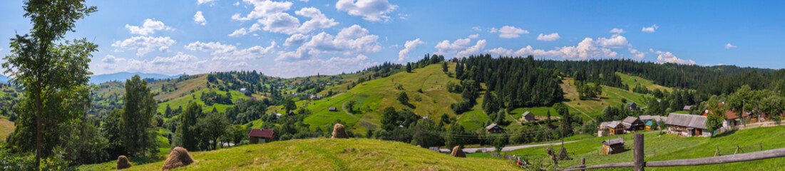 Fototapeta na wymiar A charming panorama of mountainous green slopes on a summer day with rural houses scattered around the country with dry harvested hay on farms and white rare clouds floating on the horizon line.