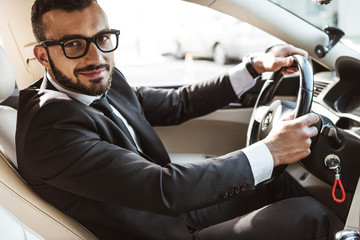 side view of handsome driver in suit driving car and looking at camera