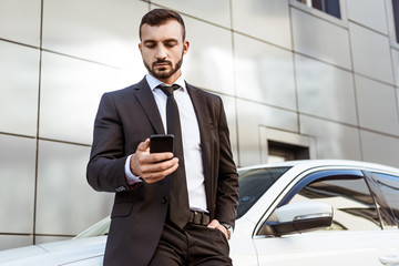 handsome businessman holding smartphone and standing near car
