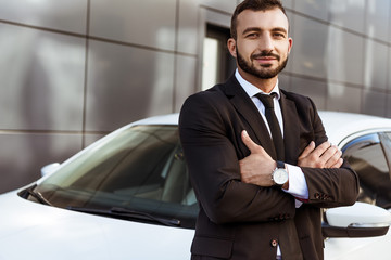 handsome smiling businessman standing with crossed arms near car