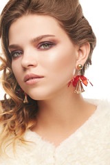 Fototapeta premium Close up portrait of a young fair-haired lady with one sided hairstyle. The girl in an ivory shaggy coat and red chunky flower earrings, posing over the white background, looking at the camera.