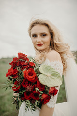 The joyful stylish beautiful fabulous happy blonde bride with the stylish bouquet is tenderly hugging flowers on the road in the countryside during the cloudy sunset.