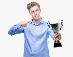 Young handsome blond man holding a trophy with surprise face pointing finger to himself