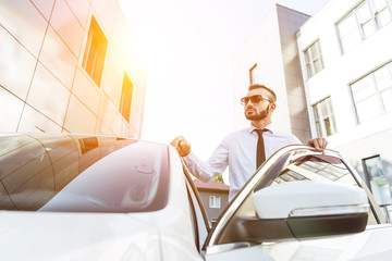 handsome driver in sunglasses standing near open car on street during sunset