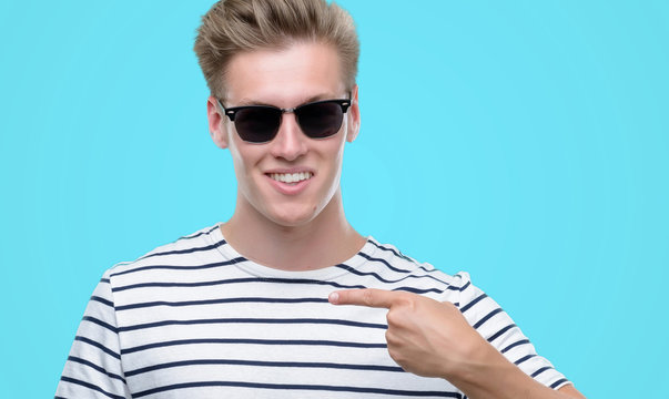 Young handsome blond man wearing sunglasess with surprise face pointing finger to himself