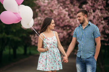 Young happy couple in love outdoors. Handsome man and beautiful woman on a walk in a spring blooming park. They keep helium balls