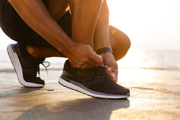 Cropped photo legs of sporty caucasian man in tracksuit and sneakers squatting on pier at seaside, and tying his shoelaces - obrazy, fototapety, plakaty