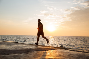 Image of muscular sporty man 30s in shorts and t-shirt running along pier at seaside, and listening to music via wireless headphones during sunrise