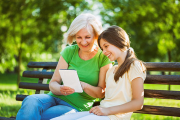 Grandmother and granddaughter are sitting in park and using digital tablet. 