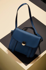 Women's blue leather handbag in vintage style, with a flap top, handle, shoulder strap and gold lock, front view. The trendy purse for office, business trips or casual occasions placed on a desk top.