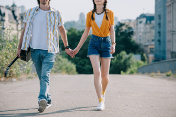 Young and happy man with guitar walking and holding hand of girl musicians in city