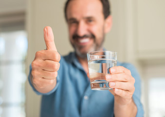 Middle age man drinking a glass of water happy with big smile doing ok sign, thumb up with fingers,...