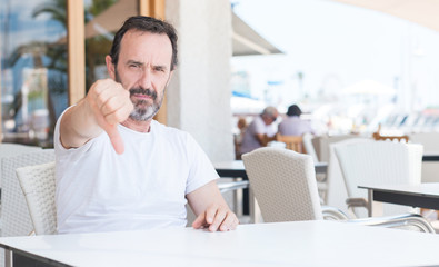 Fototapeta premium Handsome senior man sitting at terrace with angry face, negative sign showing dislike with thumbs down, rejection concept