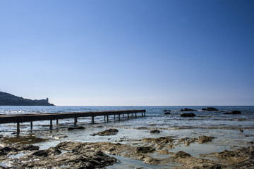 On the left side of the picture you can see the jetty. In the foreground flat rocks can be seen that look out of the water. In the background you can see a small piece of land, blue sea and blue sky.