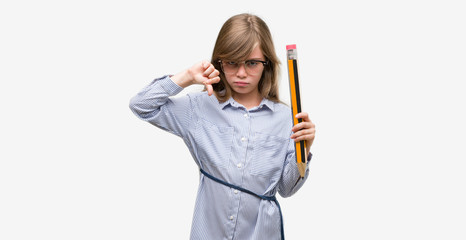 Young blonde child holding big pencil with angry face, negative sign showing dislike with thumbs down, rejection concept