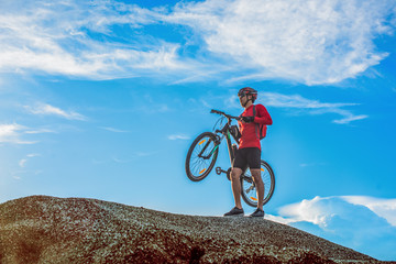 Cyclist man carrying his mountain bike along the rocky terrain on a sunny day against a blue sky