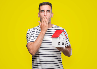 Handsome real estate agent holding a house cover mouth with hand shocked with shame for mistake,...