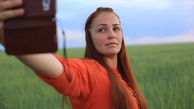 young beautiful woman in red dress and red hair, takes a selfie on the phone in the green wheat field in the evening at sunset in summer. the nature of digital technology.