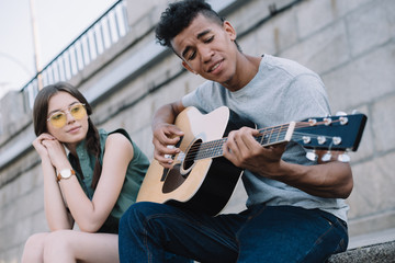 Pretty girl listening to African american man playing guitar in urban environment