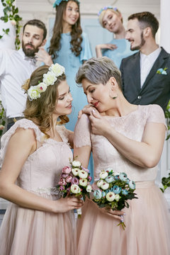 Lesbian wedding. Newlyweds in pale pink dresses posing for photos, flowers in their hands, the long-haired bride tenderly touching the shoulder of her spouse. Friends and bridesmaids standing behind.