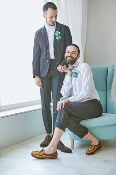 Gay Couple On Their Wedding Day. Two Happy Grooms Posing In A Luxury Hotel Room. One Guy Sitting In The Armchair, Holding Hands With His Partner, Who's Standing Beside. Both Men Wearing Boutonnieres.