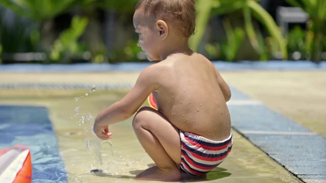 Cute toddler boy sitting in shallow water at swimming pool and playing with sprinkler at sunny summer day