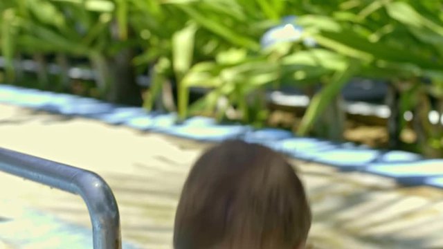 Toddler boy picking up water with plastic bucket and pouring it off in while playing in kiddie swimming pool at hot summer day