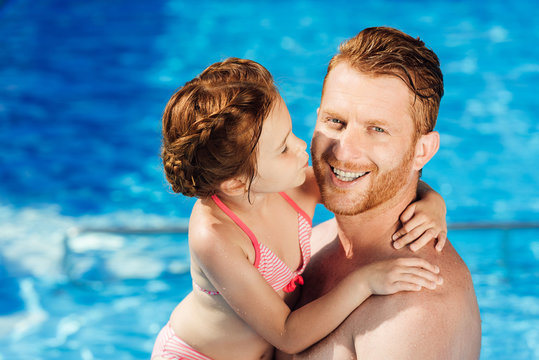 happy father and daughter embracing in swimming pool and looking at camera